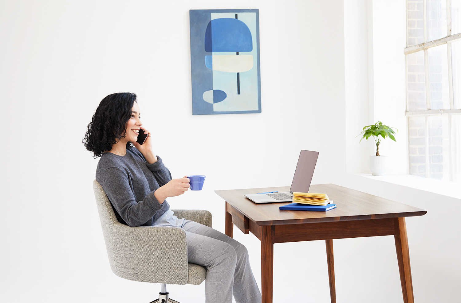 A lady speaking on her cell phone and holding a cup of coffee in front of a desk that has an open laptop