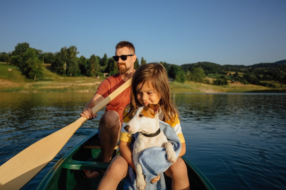 A father paddling a canoe on a lake while his daughter holds their dog wrapped in a towel