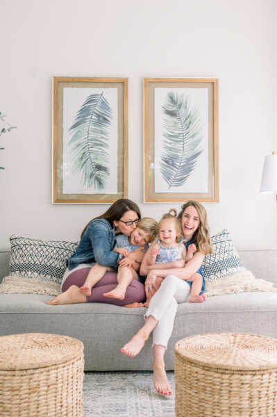 Two white women sit on a grey couch with their two children. Behind them are two portraits with leaves.