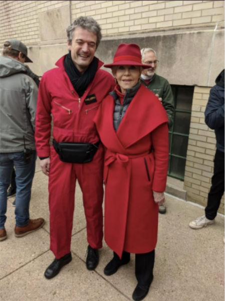 To the left, Boris Khentov is a tall white man wearing a red jumpsuit with a black scarf and black satchel around his waist. To the right, Jane Fonda is also wearing a red coat and hat, smiling.